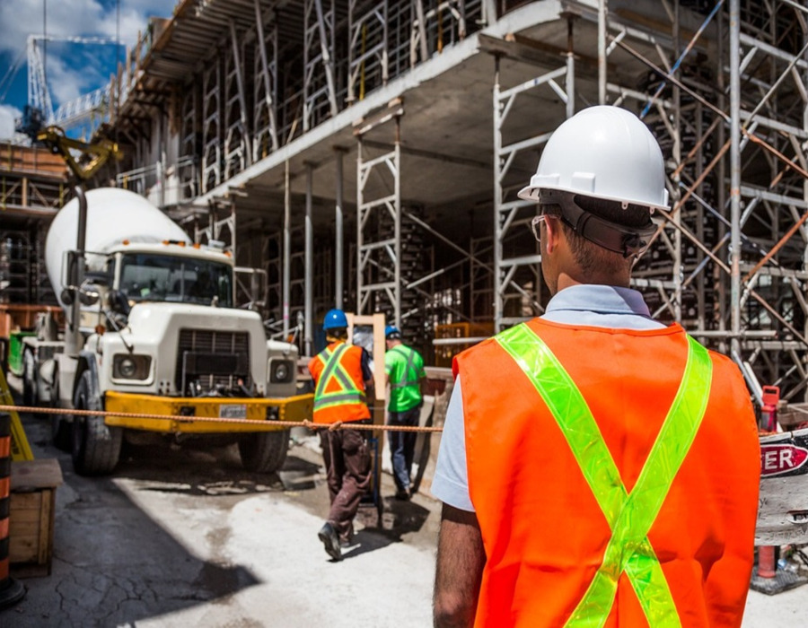 A construction worker wearing a yellow hard hat and high-visibility safety vest is focused on his work at a construction site. He is using a handheld tool, possibly a drill or a similar power tool, with concentration and precision. The background features construction materials and part of a structure under construction, highlighting the industrial environment and the theme of building and safety.